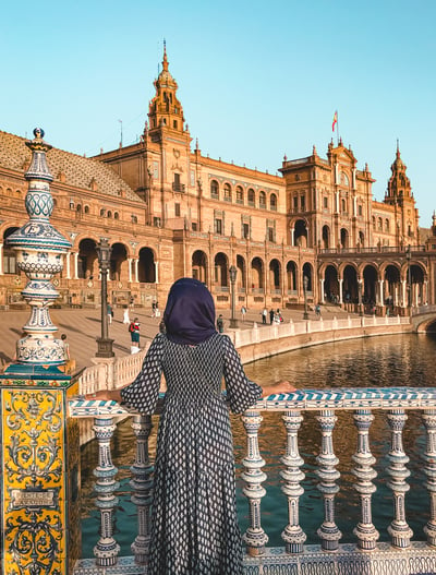 Muslim woman in blue dress standing near yellow tiled pillar on bridge at Plaza de Espana